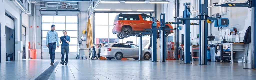 Two people walking through an automotive service workshop with cars on lifts in the background.